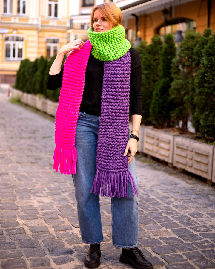 Colorful scarf with fringes in pink, green, and purple worn by a woman in a city street, showcasing winter fashion.
